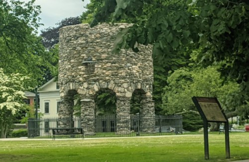 Stone tower with arched openings, surrounded by greenery and a sign nearby, set in a park-like area.
