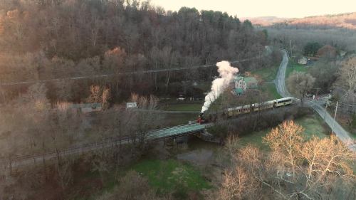 A steam train crosses a bridge surrounded by trees and hills, with smoke billowing against a sunset backdrop.