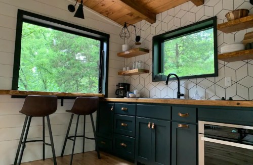 A modern kitchen with dark cabinets, wooden shelves, and large windows overlooking greenery. Two bar stools are at the counter.