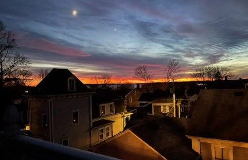 A vibrant sunset over rooftops, with colorful clouds and a crescent moon in the evening sky.