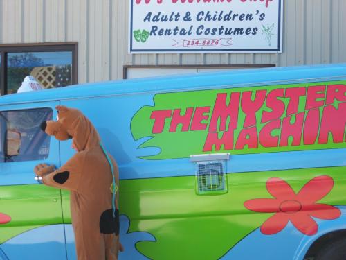 A person in a Scooby-Doo costume stands next to a colorful van labeled "The Mystery Machine."
