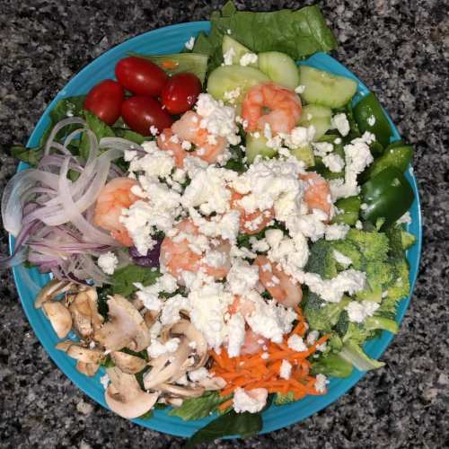 A colorful salad with shrimp, mixed greens, cherry tomatoes, cucumbers, broccoli, mushrooms, and feta cheese.