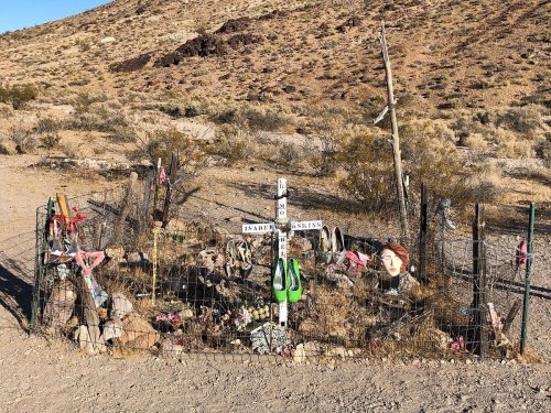 A small, fenced memorial with flowers, rocks, and decorations in a desert landscape.