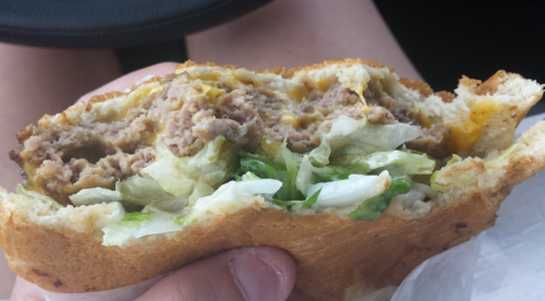 A close-up of a half-eaten sandwich with ground meat, lettuce, and cheese, held in a hand inside a car.
