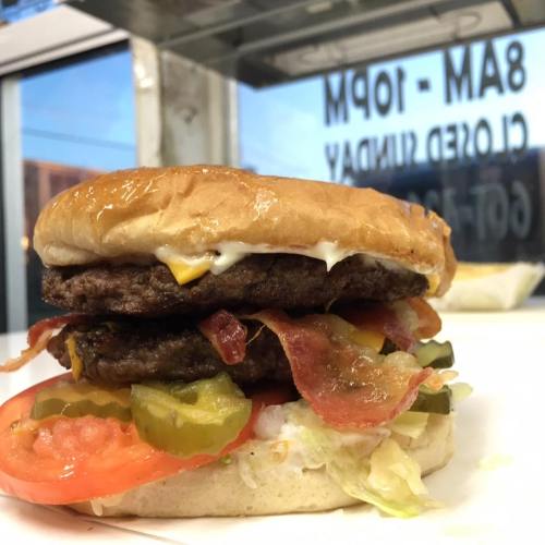 A close-up of a double cheeseburger with bacon, pickles, lettuce, and tomato, served on a soft bun.