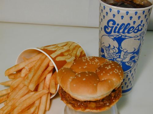 A burger with sesame seeds, a side of French fries in a cup, and a large drink in a patterned cup.