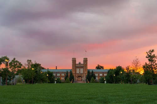 A historic brick building with towers stands against a colorful sunset, surrounded by green grass and trees.