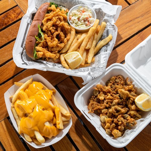 A wooden table with three food containers: a sandwich with fried seafood, fries with cheese, and fried seafood with coleslaw.