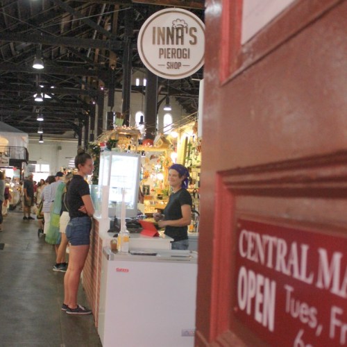 A view of a pierogi shop with a smiling employee at the counter, and customers in a bustling market setting.