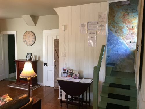 A cozy interior with a clock on the wall, a lamp, a table, and stairs leading to a colorful wall.