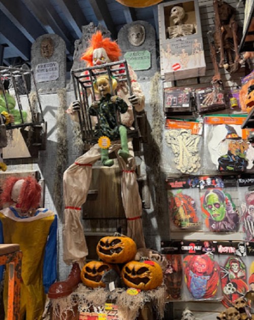 A spooky Halloween display featuring a clown in a cage, a doll, and decorated pumpkins among various creepy decorations.