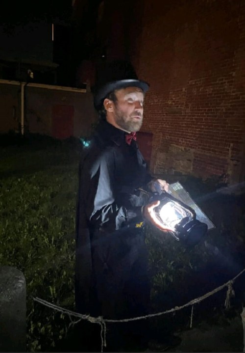 A man in a black cape and top hat holds a lantern, standing in a dimly lit outdoor area at night.