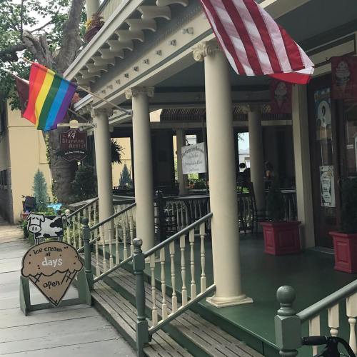 A porch with American and rainbow flags, featuring a sign that says "Open" and decorative planters.