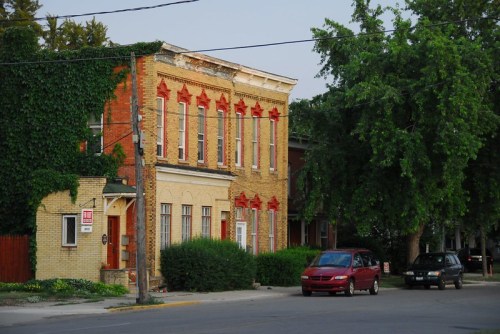 Historic brick building with red accents, surrounded by greenery and parked cars on a quiet street.