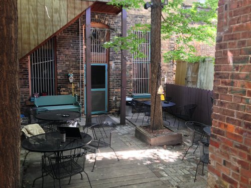 A cozy outdoor patio with tables and chairs, surrounded by brick walls and greenery.