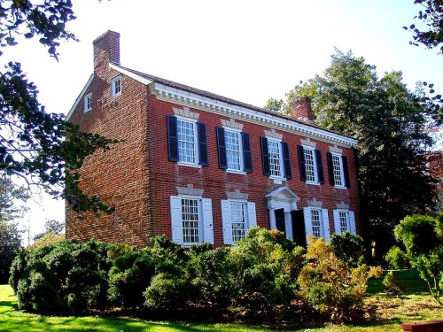 A historic brick house with white shutters and a central entrance, surrounded by greenery and trees.