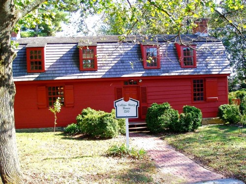 A red historic house with a shingled roof, surrounded by greenery and a sign reading "Collins Sharp House 1800."