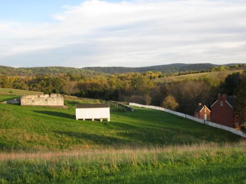 A scenic landscape featuring green fields, a stone structure, a wooden shed, and a red house against rolling hills.