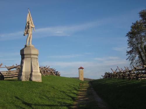 A soldier statue stands on a grassy path leading to a watchtower, flanked by wooden fences under a clear blue sky.