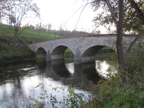A stone arch bridge spans a calm river, surrounded by trees and grassy hills. Reflections shimmer on the water's surface.