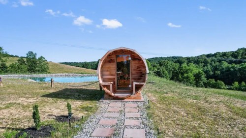 A wooden barrel-shaped structure sits on a grassy hillside near a pond, surrounded by trees and blue sky.
