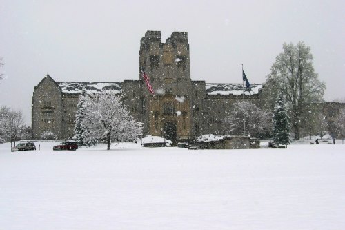 A historic stone building covered in snow, surrounded by trees and a snowy landscape, with flags flying in the foreground.