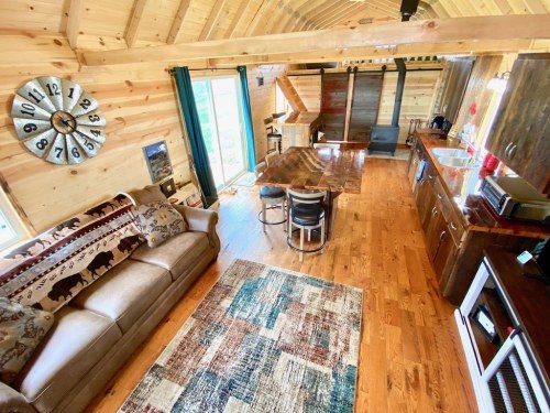 Cozy wooden cabin interior with a sofa, dining table, kitchen area, and large windows letting in natural light.