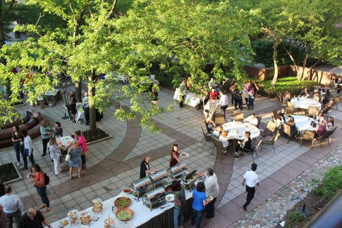 Outdoor event with guests mingling, dining at tables, and a buffet setup surrounded by greenery.