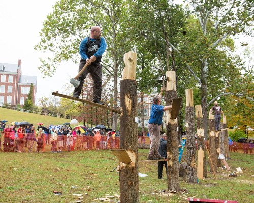 Two men chop wood on vertical logs at an outdoor event, with spectators watching under umbrellas in the background.