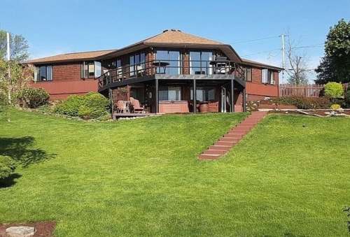 A large, modern house with a deck, surrounded by a well-maintained lawn and landscaping under a clear blue sky.