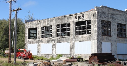Abandoned building with broken windows, surrounded by overgrown grass and old machinery. Clear blue sky above.