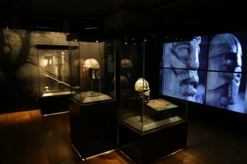 Display of ancient artifacts in a museum, featuring helmets, sculptures, and a large digital screen in the background.