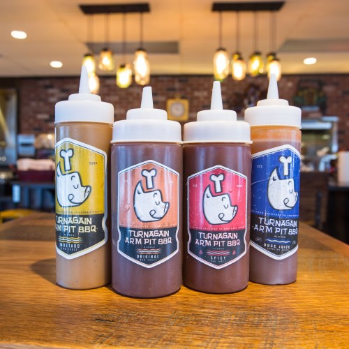 Four squeeze bottles of barbecue sauce on a wooden table, labeled Original, Spicy, and two others from Turnagain Arm Pit BBQ.