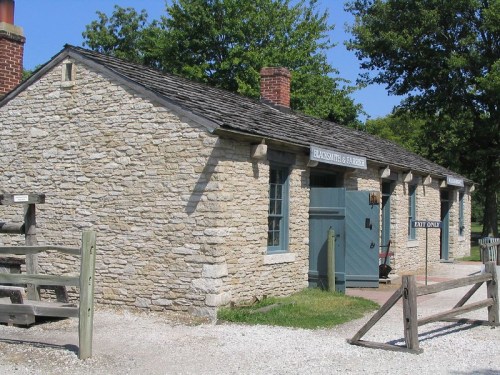 Historic stone blacksmith shop with a wooden door and sign, surrounded by greenery and a gravel path.