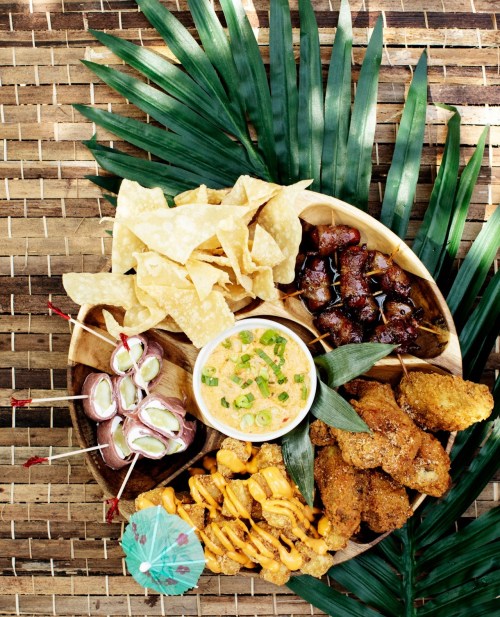 A wooden platter filled with various snacks: chips, skewers, fried chicken, and a dip, surrounded by green leaves.