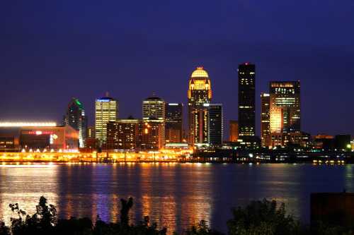 Louisville skyline at night, featuring illuminated buildings reflecting on the water.