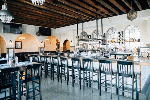 A modern restaurant interior featuring a long bar, high stools, and large windows with decorative lighting.