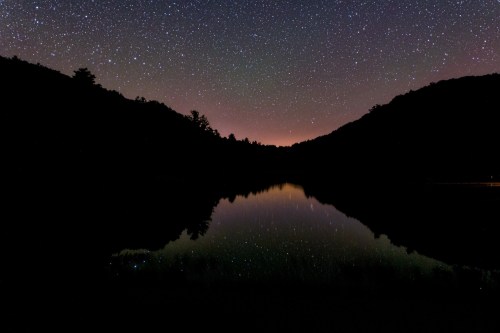 A serene night sky filled with stars reflects on a calm lake, surrounded by dark silhouettes of mountains.