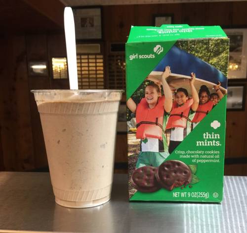 A cup of mint chocolate shake next to a box of Girl Scouts Thin Mints cookies on a wooden table.