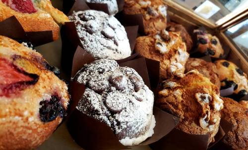A variety of freshly baked muffins in brown paper liners, featuring chocolate, fruit, and powdered sugar toppings.