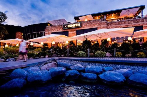 A modern restaurant with outdoor seating, surrounded by greenery and rocks, under a twilight sky.