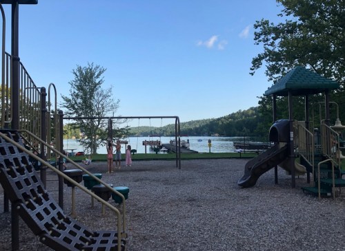 Playground with slides and swings overlooking a calm lake surrounded by trees under a clear blue sky.