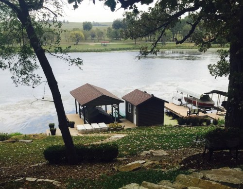 A serene lakeside view featuring a dock, two boats, and lush greenery surrounding the calm water.