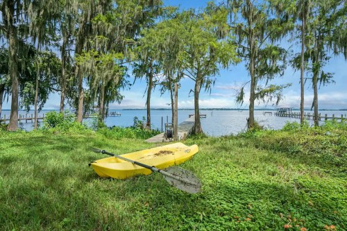 A yellow kayak rests on green grass by a calm lake, surrounded by trees and a wooden dock in the background.