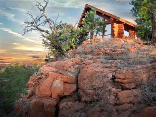 A rustic cabin perched on a rocky hillside, surrounded by trees and a colorful sunset sky.