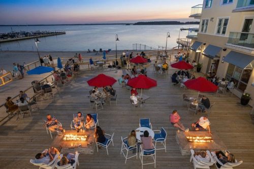 A waterfront patio with people gathered around fire pits, red umbrellas, and a sunset over the water in the background.