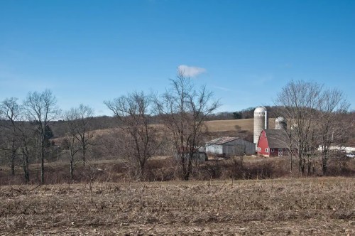 A rural landscape featuring a red barn and silos, surrounded by bare trees and open fields under a clear blue sky.