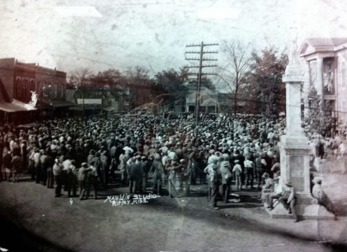 A large crowd gathers in a town square, with buildings and a monument visible in the background.