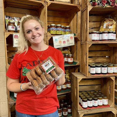 A smiling woman in a red shirt holds a tray of sausages in front of wooden crates filled with jars and products.