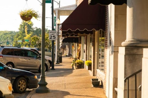 A sunny street scene with shops, parked cars, and hanging flower baskets along the sidewalk.